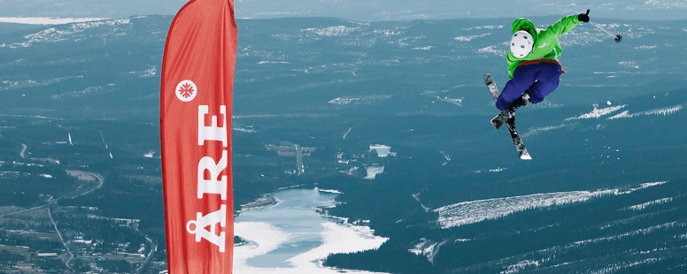 Ski in Åre A skier mid-air doing tricks. Background is a snowy forest landscape. A red banner is seen which reads Åre.